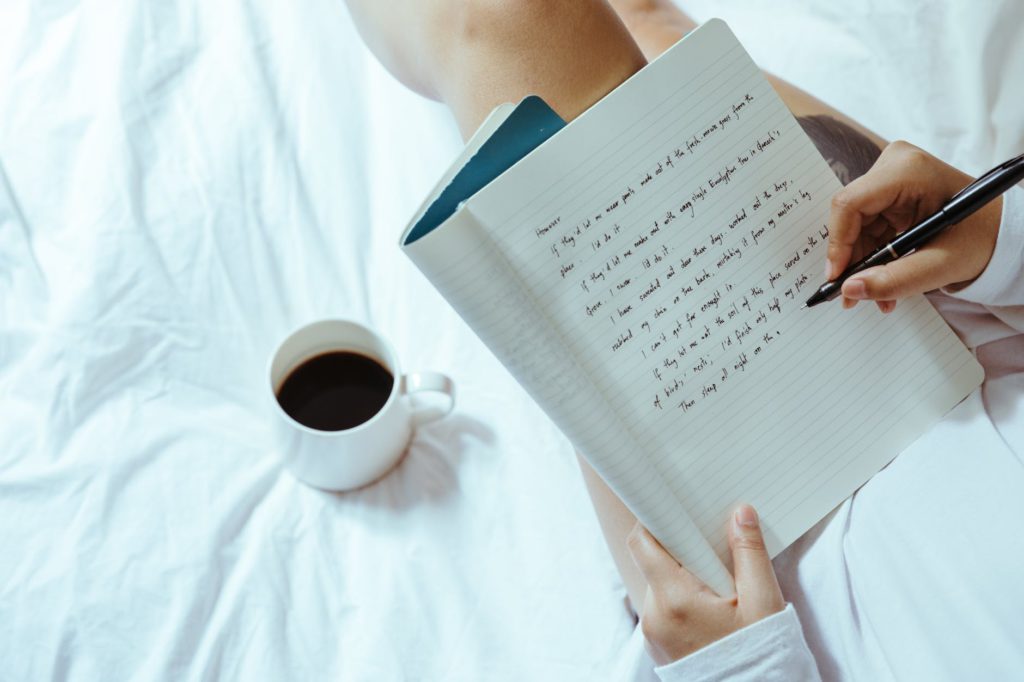 crop woman with coffee writing in notebook on bed